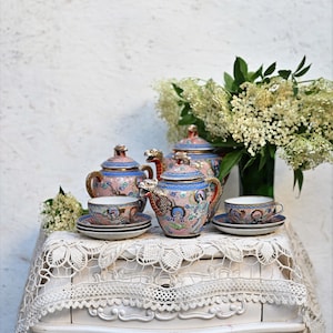 Op de afbeelding: Een vintage theeservies met een bloemenmotief in tinten blauw, roze en goud. De set bevat een theepot, roomkan, suikerpot en twee kopjes en schoteltjes. Het theeservies wordt getoond op een wit kantkleed op een witte tafel.