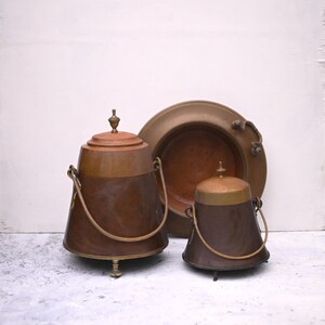 May include: Three antique copper and brass containers with lids and handles. The largest container has three legs and a knob on the lid. The smallest container has a lid with a knob and a handle. The middle container is a shallow bowl with a handle.