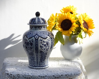 May include: A white ceramic vase with blue and white floral designs. The vase has a lid and is decorated with intricate patterns. A bouquet of yellow sunflowers is in the vase.