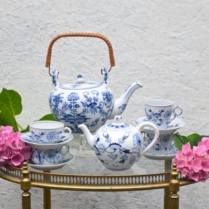 Puede incluir: Un juego de té de porcelana blanca y azul con motivos florales, que incluye una tetera con asa trenzada, una tetera más pequeña y tazas con platillos. El juego se exhibe en un carrito dorado, con flores rosas a ambos lados.