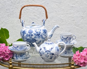 Pode incluir: Um conjunto de chá de porcelana branca e azul com padrões florais, incluindo uma chaleira com uma alça tecida, uma chaleira menor e xícaras com pires. O conjunto é exibido em um carrinho dourado, com flores cor-de-rosa de cada lado.
