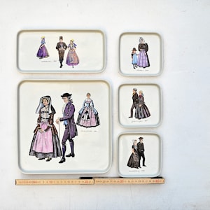 May include: Five white ceramic trays with images of people in traditional clothing. The trays are arranged in a vertical row. The top tray has two women and a man, the second tray has a man and a woman, the third tray has a man and a woman, the fourth tray has a man and a woman, and the bottom tray has a man and a woman.