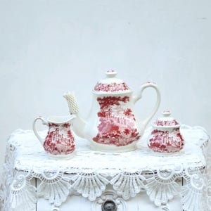 May include: A white ceramic tea set with a red landscape design. The set includes a teapot, creamer, and sugar bowl. The teapot has a textured spout and handle. The creamer and sugar bowl have matching lids. The set is displayed on a white lace tablecloth.