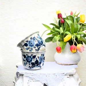 May include: Two blue and white Delftware ceramic planters with floral designs, sitting on a white lace doily on a white painted table with a bouquet of yellow and red tulips in a white vase.