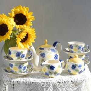 Op de afbeelding: Een bloemig theeservies met een theepot, roomkan, suikerpot en vier kopjes en schoteltjes. De set is versierd met blauwe en gele bloemen op een witte achtergrond. Het theeservies staat op een wit kantkleed met een boeket zonnebloemen op de achtergrond.