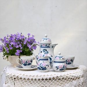 May include: A white tea set with blue and pink floral designs. The set includes a teapot, creamer, sugar bowl, and a teacup and saucer. The tea set is on a white crocheted tablecloth.