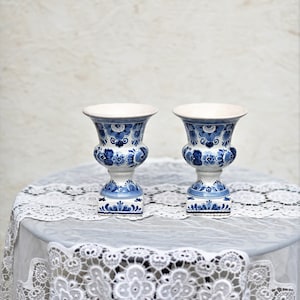 May include: Two blue and white Delftware ceramic vases with floral designs. The vases are on a white lace tablecloth.
