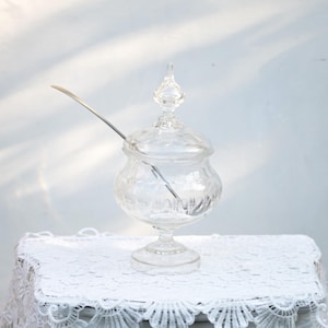 Op de afbeelding: Een heldere glazen snoepschaal met een deksel en een zilveren lepel. De schaal staat op een witte tafel met een wit kanten tafelkleed. De schaal heeft een decoratief patroon en een voet.