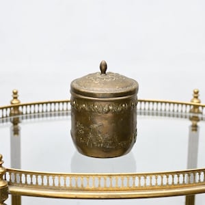 May include: An antique brass container with a lid. The container has a detailed landscape scene with a windmill. The lid has a small knob and decorative patterns. The container is on a glass surface.