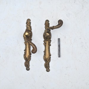 Op de afbeelding: Twee sierlijke gouden deurgrepen met ingewikkelde details. De grepen bevinden zich op een witte achtergrond.