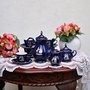 May include: A blue and gold tea set with a teapot, creamer, sugar bowl, and two cups and saucers. The set is decorated with a floral pattern and is sitting on a lace tablecloth.