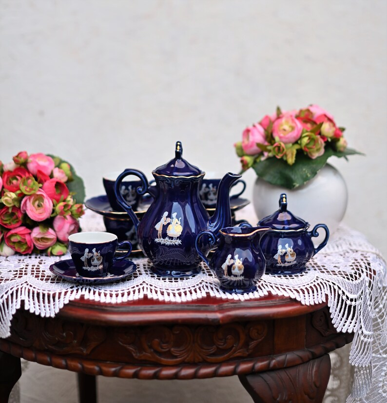 May include: A blue and gold tea set with a teapot, creamer, sugar bowl, and two cups and saucers. The set is decorated with a floral pattern and is sitting on a lace tablecloth.