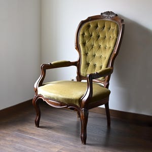 Peut inclure: Fauteuil ancien avec un dossier haut et un cadre en bois courbé. Le fauteuil présente une assise et un dossier en velours vert olive capitonné. Le cadre en bois comporte des sculptures et des accoudoirs, mettant en valeur un design classique.