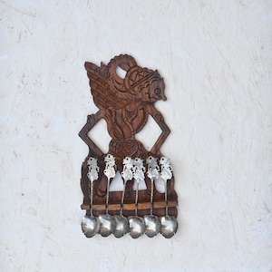Puede incluir: Un soporte de pared de madera para cucharas tallado en forma de criatura mítica con alas. El soporte tiene ocho cucharas de plata colgando.