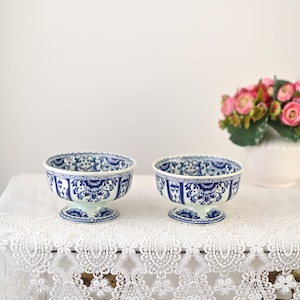 May include: Two white ceramic bowls with blue and white floral designs. The bowls have a pedestal base and are sitting on a white lace tablecloth.