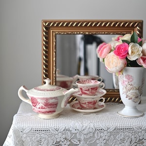 May include: A vintage tea set with a teapot, cups, and saucers in a pink and white floral pattern. The set is displayed on a white lace tablecloth with a vase of pink flowers and a gold framed mirror in the background.