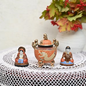 May include: A set of three ceramic figurines, including a red and gold incense burner with a dragon design, and two smaller figurines of people in traditional Japanese clothing. The figurines are on a white crocheted table cloth.