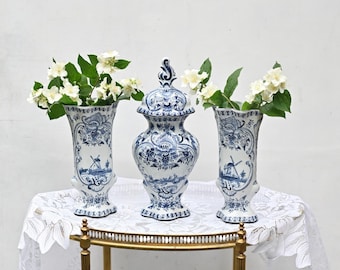 May include: Three blue and white Delftware vases with floral designs and windmill motifs. The vases are arranged on a gold metal stand with a white lace tablecloth.