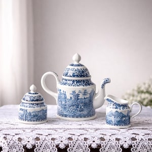 Puede incluir: Un juego de té de porcelana con un diseño de paisaje azul y blanco. El juego incluye una tetera, un azucarero y una jarra de crema. El juego de té se exhibe sobre un mantel de encaje blanco.