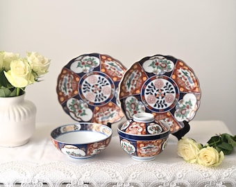 Può includere: Un set di stoviglie in porcellana giapponese, tra cui piatti, ciotole e un piatto da portata con coperchio. I pezzi hanno un bordo smerlato e sono decorati con motivi floreali e geometrici nei toni del rosso, blu e bianco.