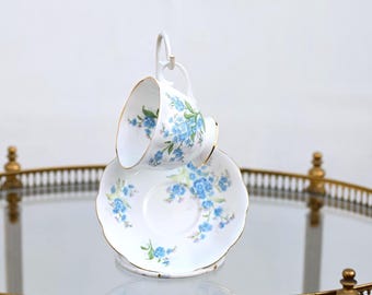 May include: A white teacup and saucer set with a delicate blue floral pattern and gold trim. The teacup is suspended above the saucer on a white stand, showcasing the design. The set is displayed on a glass-topped, gold-framed serving cart.