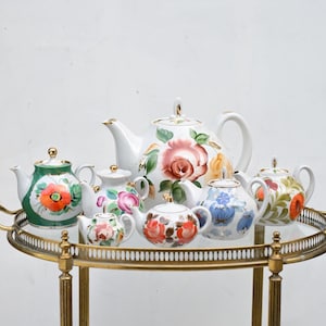 May include: A collection of decorative teapots with floral designs. The teapots are white, green, and blue, with gold accents. They are arranged on a gold-colored serving cart with a glass top.