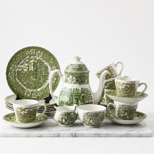 Può includere: Un servizio da tè in ceramica verde e bianca con un motivo a paesaggio. Il set comprende una teiera, tazze, piattini, piatti e una lattiera. I piatti e le tazze sono impilati e disposti su una superficie bianca.