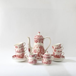 Può includere: Un servizio da tè vintage con un motivo floreale rosso e bianco. Il set include una teiera, una lattiera, una zuccheriera, piattini e tazze da tè. La teiera è alta circa 25 cm. Le tazze sono impilate a coppie.