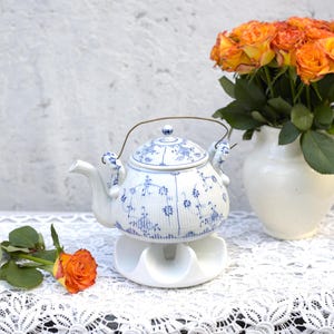 Puede incluir: Una tetera blanca con diseños florales azules y un soporte blanco sobre un mantel de encaje blanco. Un ramo de rosas naranjas en un jarrón blanco está en el fondo.