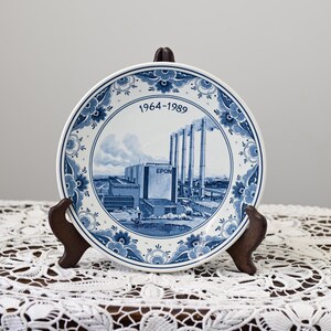 Könnte beinhalten: Ein dekorativer blau-weißer Teller mit einer Fabrikszene mit dem Text "EPON" und "hunzecentrale". Der Teller hat einen floralen Rand und die Jahreszahlen "1964-1989". Er wird auf einem Holzständer präsentiert.