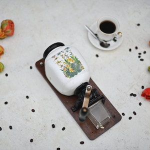 Op de afbeelding: Een vintage witte koffiemolen met een bloemenontwerp, gemonteerd op een houten basis, naast een kopje koffie en verspreide koffiebonen. Oranje en gele tulpen voegen kleur toe.