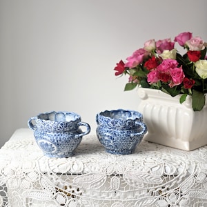 May include: Two blue and white ceramic planters with handles, sitting on a white lace tablecloth. A white planter with pink and white flowers is in the background.