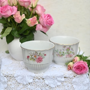 May include: Two white ceramic flower pots with floral designs on a white crocheted doily. The pots are on a white table with pink roses in the background.