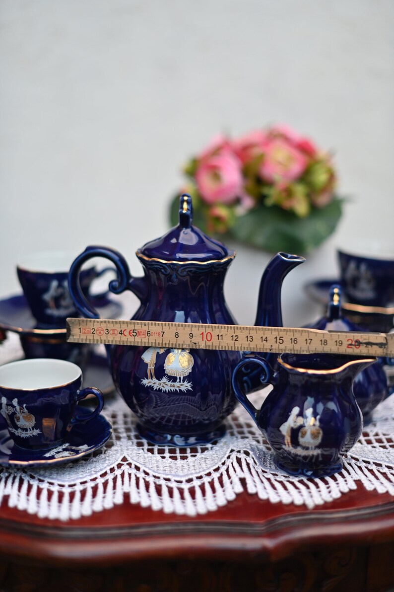 May include: A blue and gold tea set with a teapot, creamer, sugar bowl, and two cups and saucers. The set features a delicate floral design and is displayed on a white lace tablecloth.