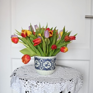 May include: A vibrant bouquet of tulips in a blue and white patterned ceramic pot. The tulips display a mix of red, yellow, and purple blooms. The pot sits on a small, round table covered with a white lace doily.