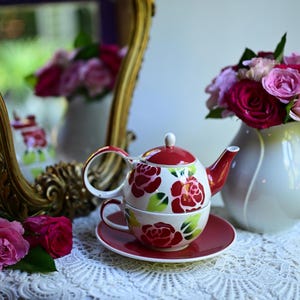 Peut inclure: Un ensemble théier et tasse en céramique rouge et blanc avec un motif floral de roses. L'ensemble est posé sur une nappe en dentelle blanche.