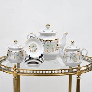 May include: A vintage tea set with a white base, gold trim, and leaf pattern. The set includes a teapot, creamer, sugar bowl, and small saucers. The tea set is displayed on a gold-colored serving cart.