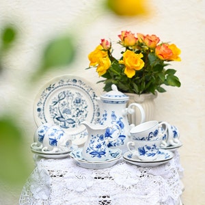 Peut inclure: Un service à thé en porcelaine blanche avec des motifs floraux bleus, comprenant une théière, un pot à crème, un sucrier et des tasses et soucoupes. L'ensemble est disposé sur une nappe en crochet blanche sur une table blanche. Un bouquet de roses jaunes et orange est en arrière-plan.