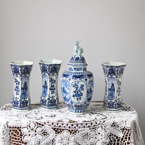May include: Four blue and white ceramic vases and a lidded jar with a dog figurine on top. The vases and jar are decorated with floral and windmill motifs. They are arranged on a table covered with a white lace tablecloth.