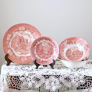 Puede incluir: Un conjunto de platos y un cuenco de cerámica rosa y blanca antiguos con un diseño paisajístico. Los platos se exhiben sobre pequeños soportes negros, y el cuenco descansa en otro cuenco blanco. Los artículos están dispuestos sobre un mantel de encaje blanco.