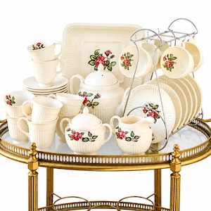 May include: A cream-colored ceramic tea set featuring a red rose and green leaf pattern. The set includes a teapot, sugar bowl, creamer, multiple cups, saucers, and a square plate, with several pieces organized on a chrome wire rack stand.
