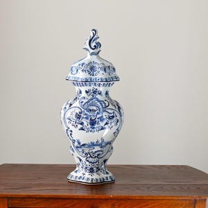 May include: A blue and white ceramic vase with a lid, featuring intricate floral and landscape patterns. The vase is displayed on a wooden surface. The design is reminiscent of Delftware.