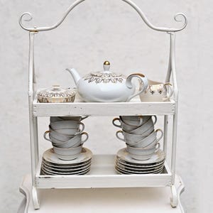 May include: White two-tiered metal serving stand with a teapot, sugar bowl, and cups and saucers. The stand has a white painted finish and ornate scrollwork details.