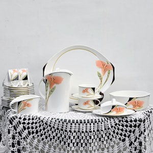 May include: A white porcelain dinner service with a floral pattern in orange and green. The set includes plates, cups, saucers, a jug, and a serving bowl. The items are displayed on a table covered with a white lace tablecloth.