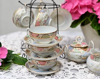May include: A vintage tea set with floral and gold accents. The set includes teacups, saucers, a teapot, and a creamer, arranged on a white lace tablecloth. A black metal stand holds additional teacups. Pink flowers add a pop of color.