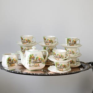 May include: A vintage tea set with a village scene design. The set includes a teapot, creamer, sugar bowl, and several teacups and saucers. The cups and saucers are stacked on a silver tray. The set has a cream background with green trim.