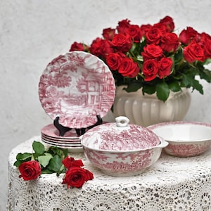Könnte beinhalten: Ein Set aus rosa und weiß gemustertem Porzellan, bestehend aus einer Schüssel mit Deckel, einer kleineren Schüssel und einem Stapel Teller. Das Geschirr ist auf einer weißen Spitzentischdecke arrangiert, im Hintergrund ein Strauß roter Rosen in einer weißen Vase.