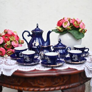 May include: A blue and white porcelain tea set with a teapot, creamer, sugar bowl, and four teacups and saucers. The set is decorated with a floral pattern and gold accents. The tea set is displayed on a wooden table with a white lace tablecloth.