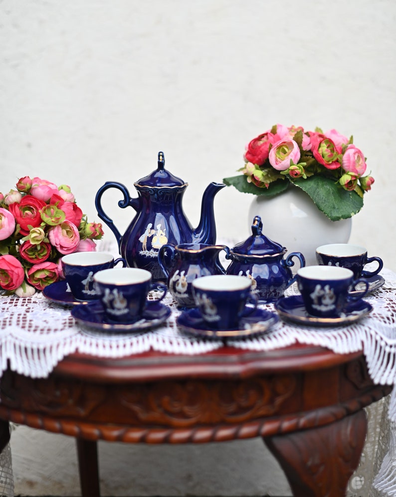 May include: A blue and white porcelain tea set with a teapot, creamer, sugar bowl, and four teacups and saucers. The set is decorated with a floral pattern and gold accents. The tea set is displayed on a wooden table with a white lace tablecloth.