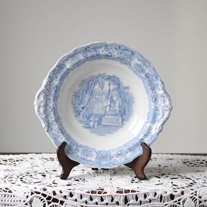 May include: A blue and white antique porcelain plate with a scalloped edge, displayed on a wooden stand. The plate features a detailed landscape scene in the center and decorative border. The plate is set on a white lace tablecloth.